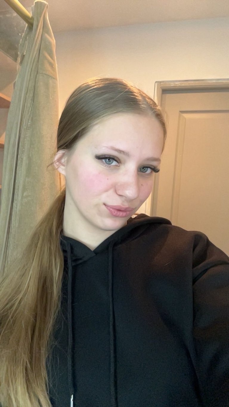 Young woman with long reddish-brown hair wearing a black hoodie, posing for a selfie indoors near a doorway