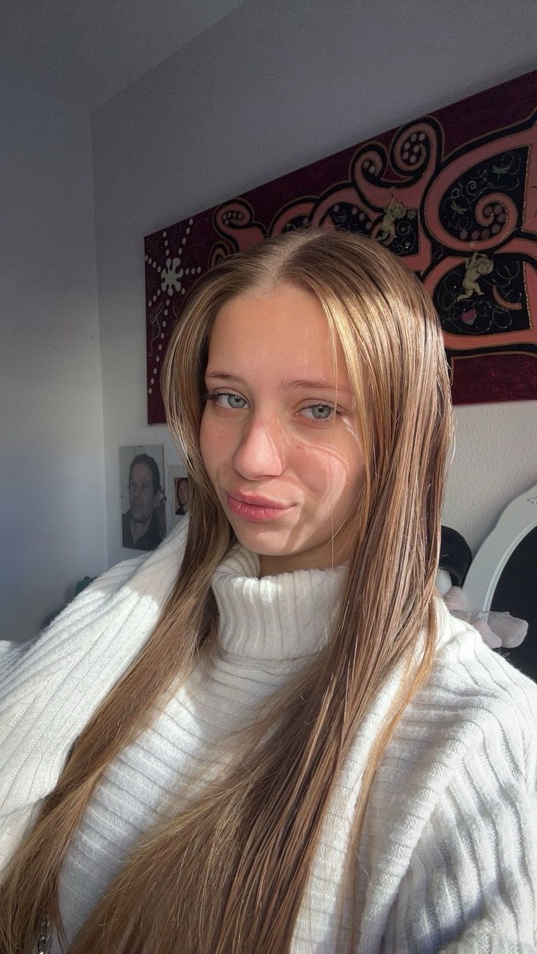 Young woman with long blonde hair wearing a white ribbed turtleneck, smiling at camera in front of decorative wall tapestry
